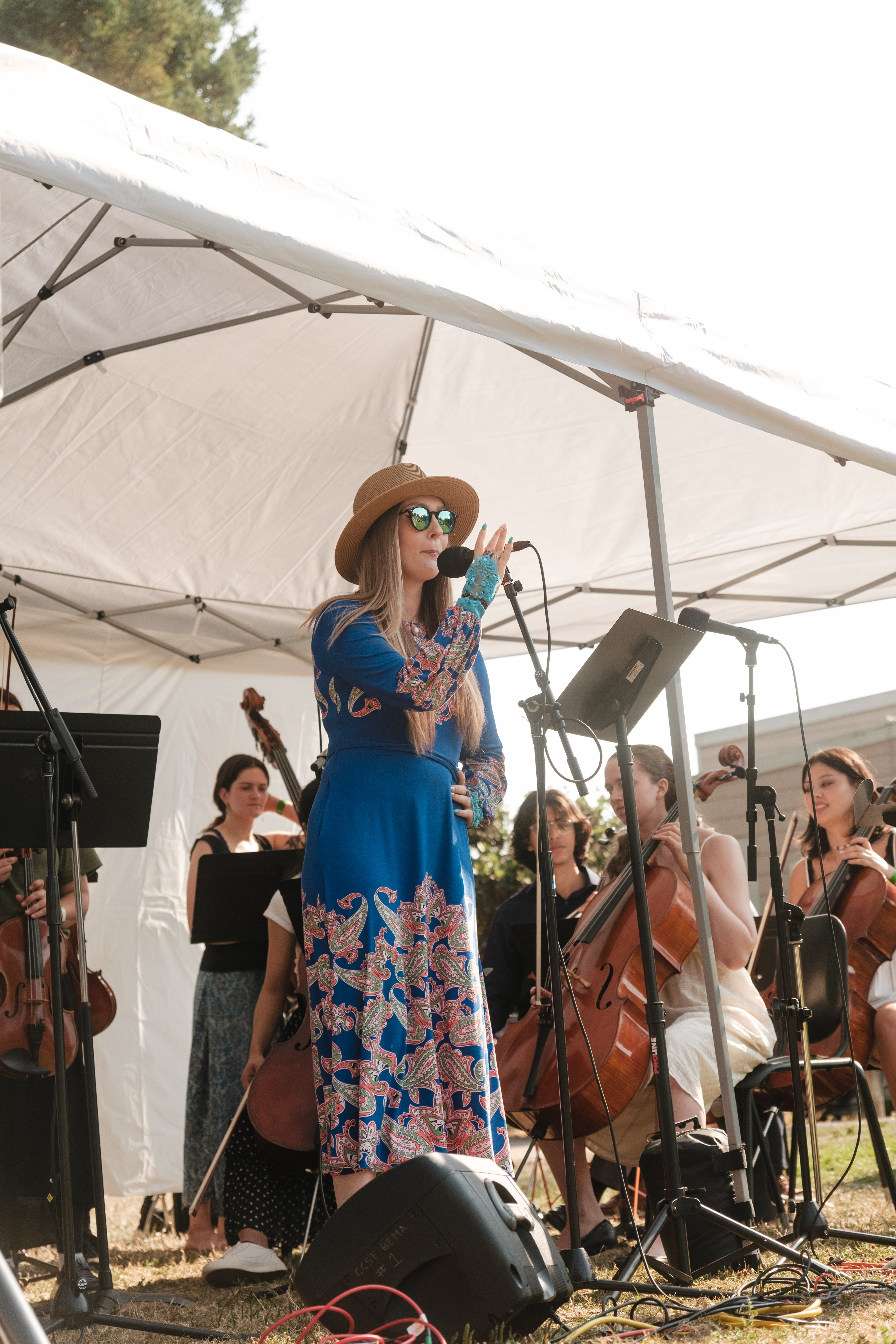 A blonde-haired woman in concert attire stands at a microphone while performing with a classical group under a white tent.